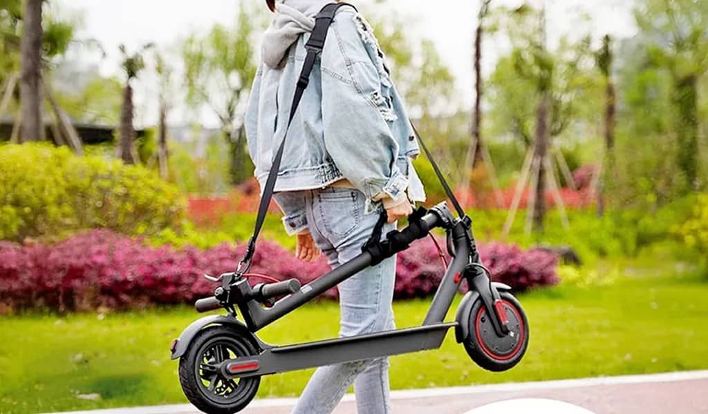 A woman carrying an electric scooter using both a shoulder straip and addon grip handle